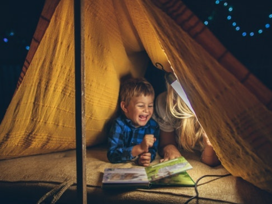 An adult and child reading inside a tent beneath a hanging light.